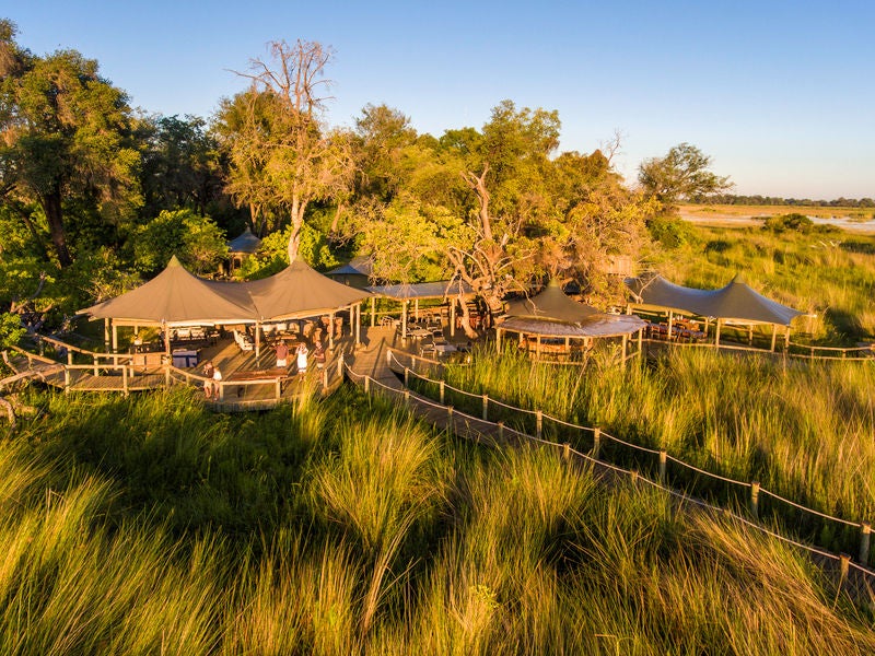 Elevated safari lodge with thatched-roof villas perched on wooden platforms overlooking Okavango Delta's pristine waterways at sunset