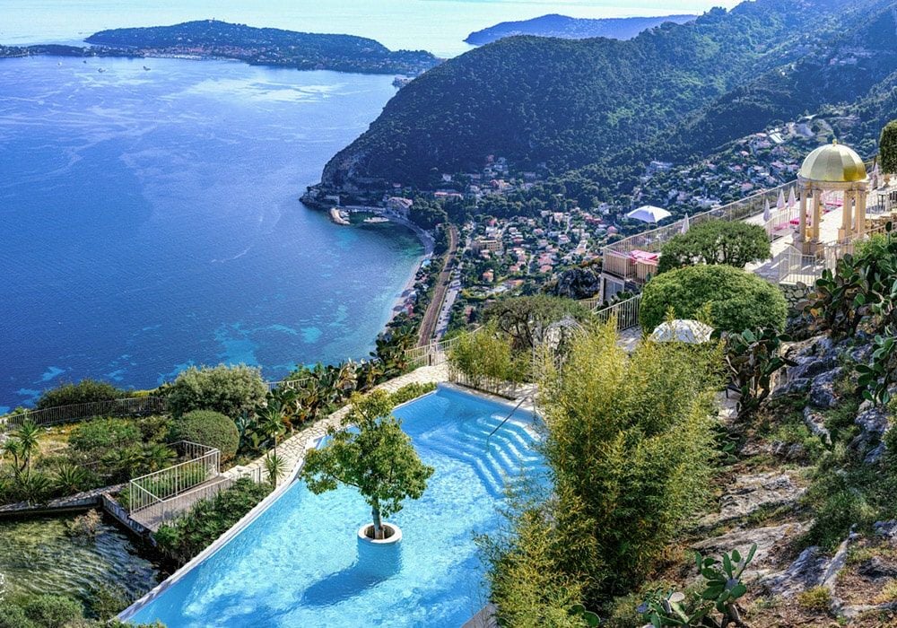 Medieval cliffside luxury hotel overlooking French Riviera, with golden stone walls and terraced gardens against Mediterranean views