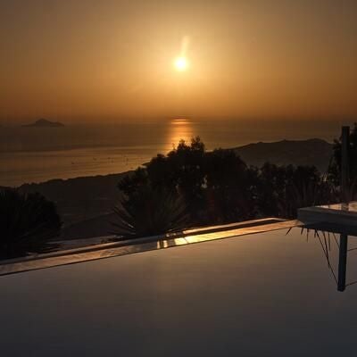 Luxurious white-washed hotel nestled on Santorini's cliffside, featuring blue-domed roofs, infinity pool overlooking deep blue Aegean Sea at sunset