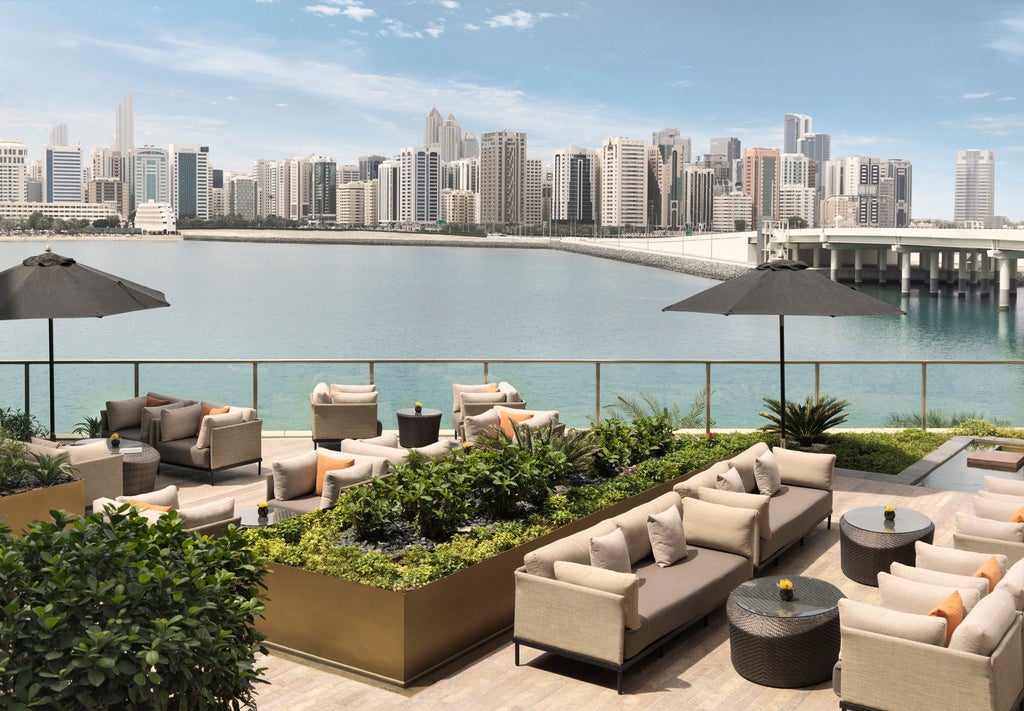 Modern luxury hotel with curved glass facade reflecting sunset, waterfront infinity pool, and Abu Dhabi skyline in background