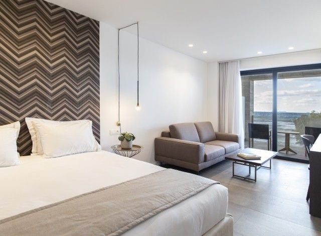 Modern junior suite with panoramic Aegean Sea view, minimalist white decor, king bed, sleek furniture, and expansive glass balcony at luxurious Greek island hotel