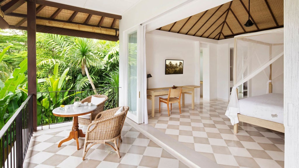 Luxurious Balinese-style room with king bed, white linens, dark wood furnishings, traditional artwork and floor-to-ceiling windows overlooking garden