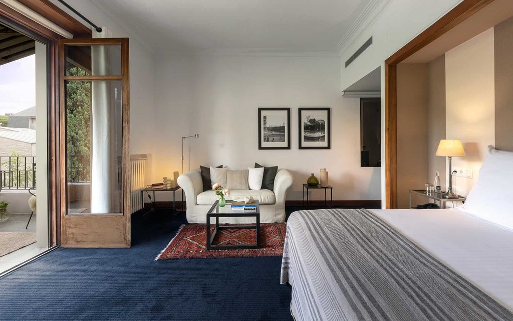 Elegant junior suite with modern Spanish design, plush white bedding, sleek wood furnishings, and expansive city-view windows at luxury hotel