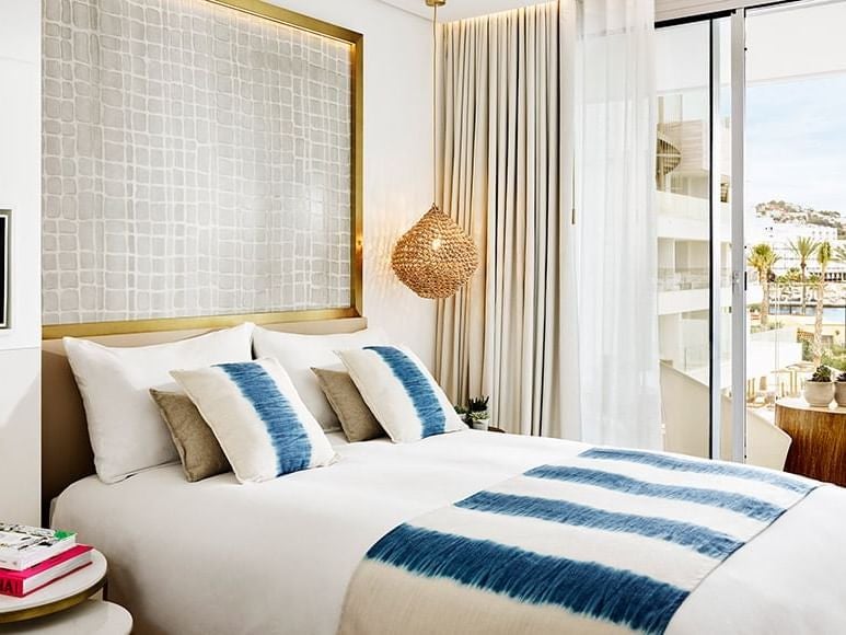 Luxurious white-themed deluxe room at Nobu Hotel Ibiza Bay with modern design, plush bedding, and panoramic coastal view through large windows.