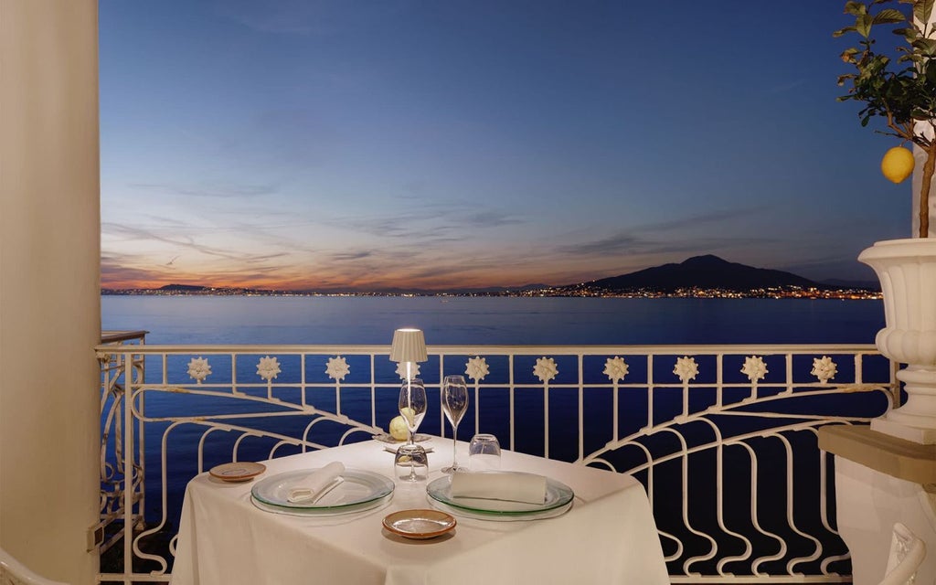 Luxurious seaside hotel with elegant white facade overlooking azure Mediterranean waters, nestled dramatically on rocky Sorrento coastline at sunset