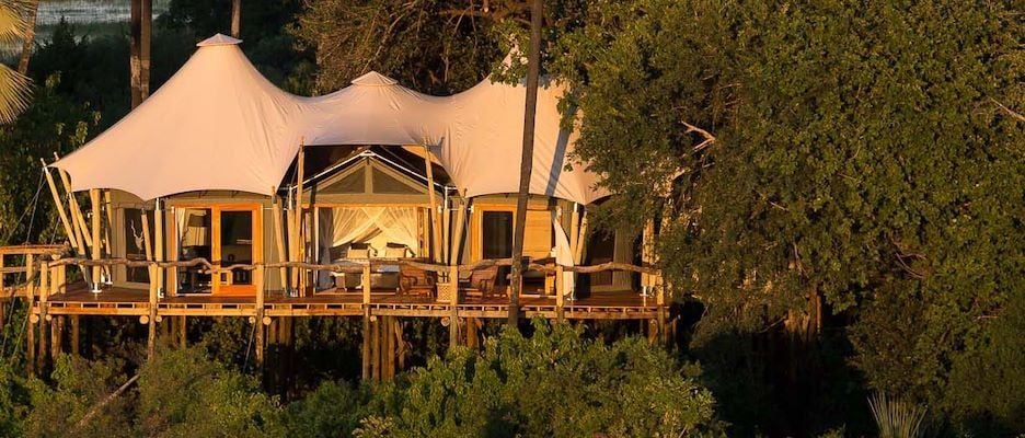 Luxurious safari tents nestled among acacia trees, overlooking a serene floodplain in the Okavango Delta, with warm golden light and wildlife silhouettes