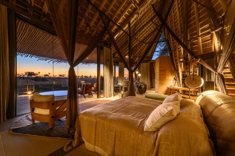 Elevated safari lodge in Botswana with thatched-roof suites on stilts above wetlands, surrounded by lush palm trees at sunset