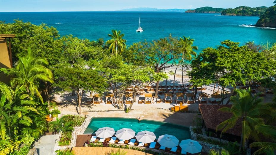 Panoramic view of luxury beachfront resort with infinity pool overlooking Pacific Ocean, surrounded by lush tropical gardens at sunset