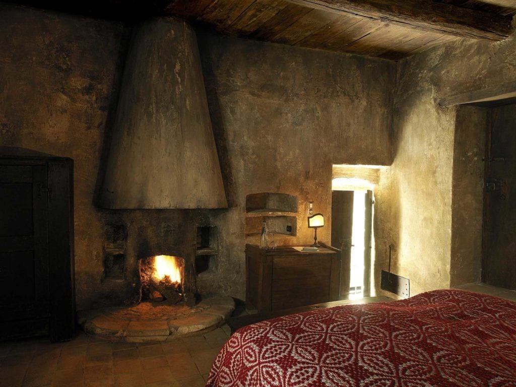 Historic Italian stone room with rustic wooden bed, arched window overlooking medieval village rooftops, warm natural light, traditional Abruzzo mountain hospitality design