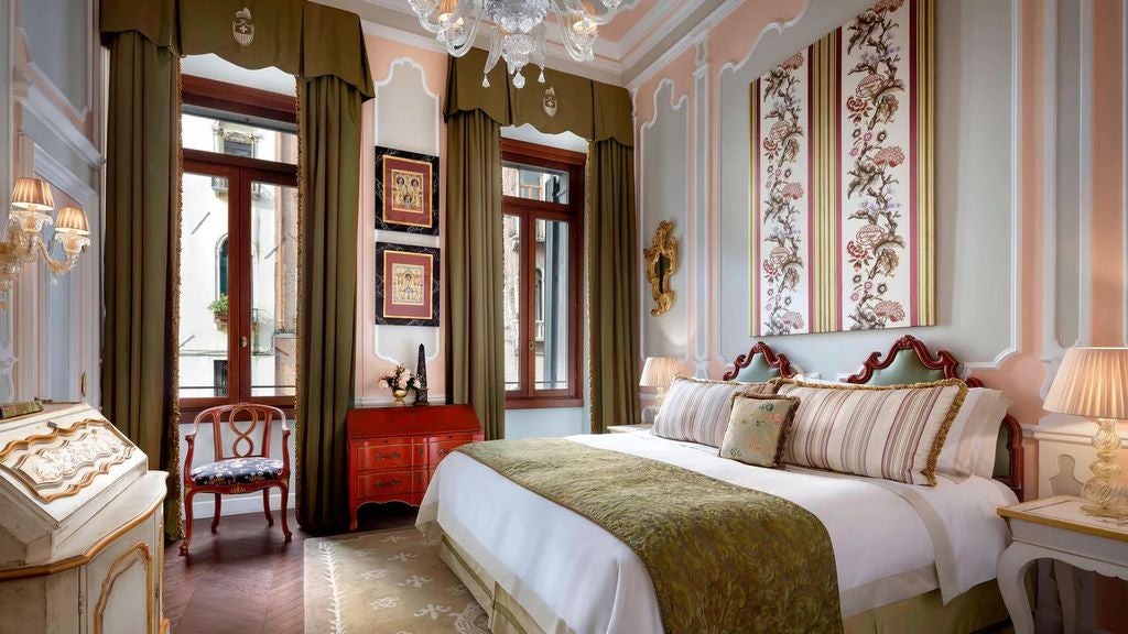 Opulent hotel room with ornate gold-framed Venetian mirrors, crystal chandelier, antique furnishings and silk-draped windows overlooking canal