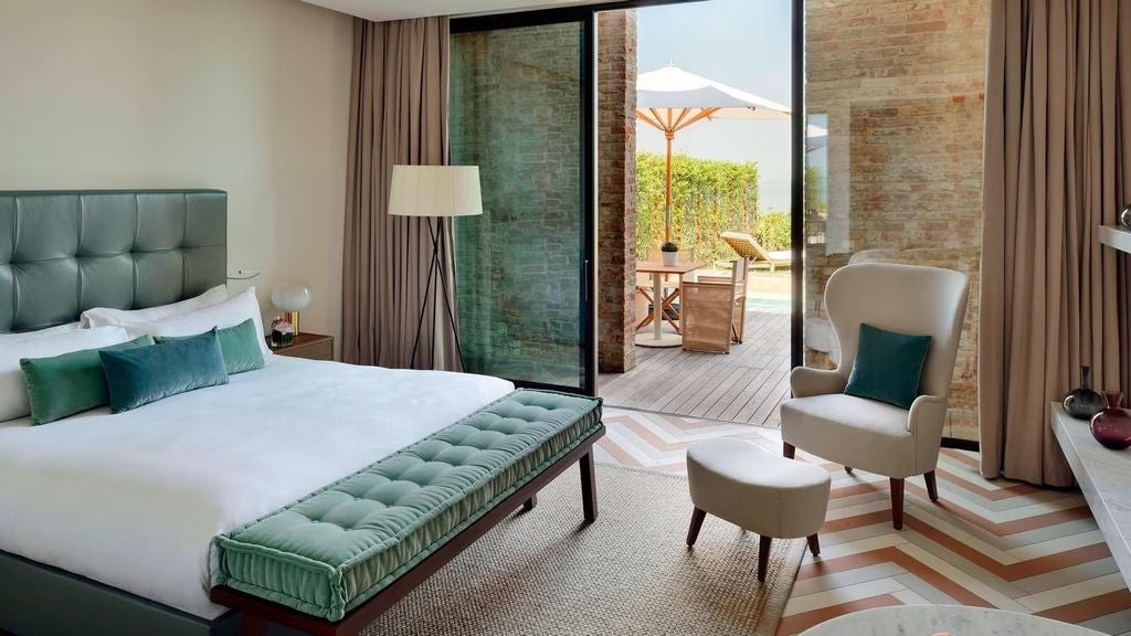 Elegant hotel room with floor-to-ceiling windows, king bed, modern decor, and private pool terrace overlooking Venice lagoon at sunset