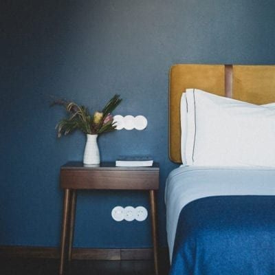 Elegant vintage-style hotel room with plush white bedding, soft neutral tones, decorative wooden headboard, and large windows overlooking Portuguese cityscape