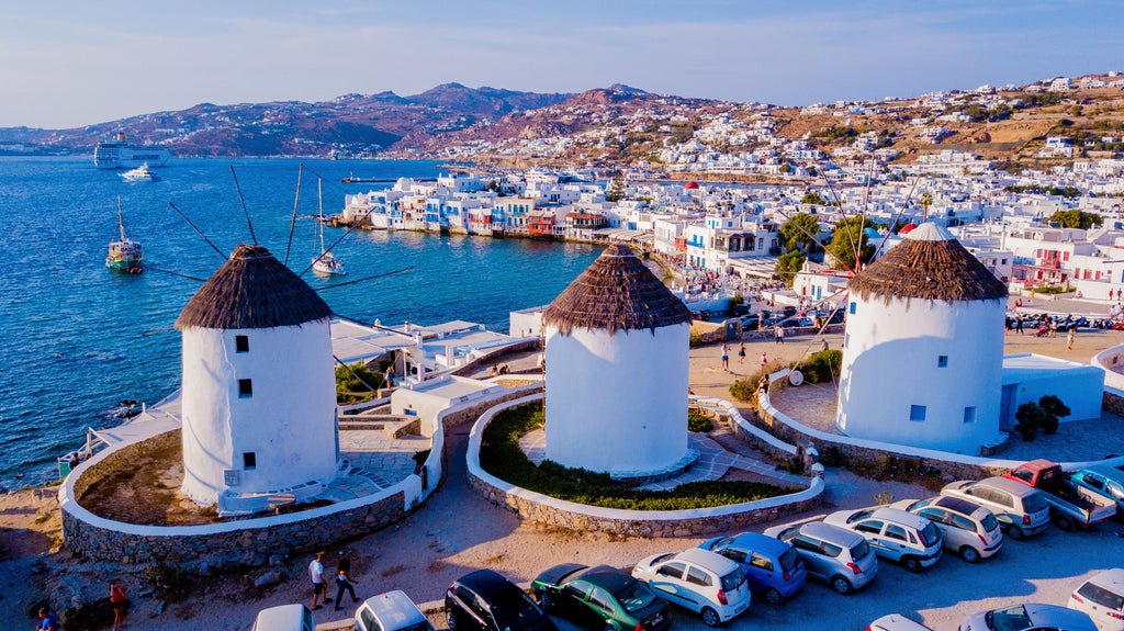 Aerial view of Mykonos harbor with pristine white buildings, luxury yachts dotting turquoise waters, and iconic windmills at sunset