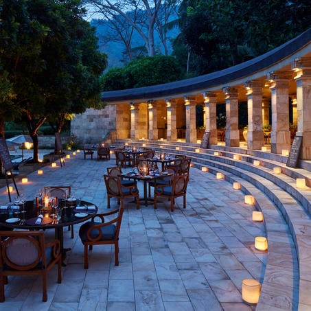 Luxury Javanese hotel with colonnaded walkway and stone rotunda, framed by lush jungle and Mount Merapi's misty silhouette at sunset