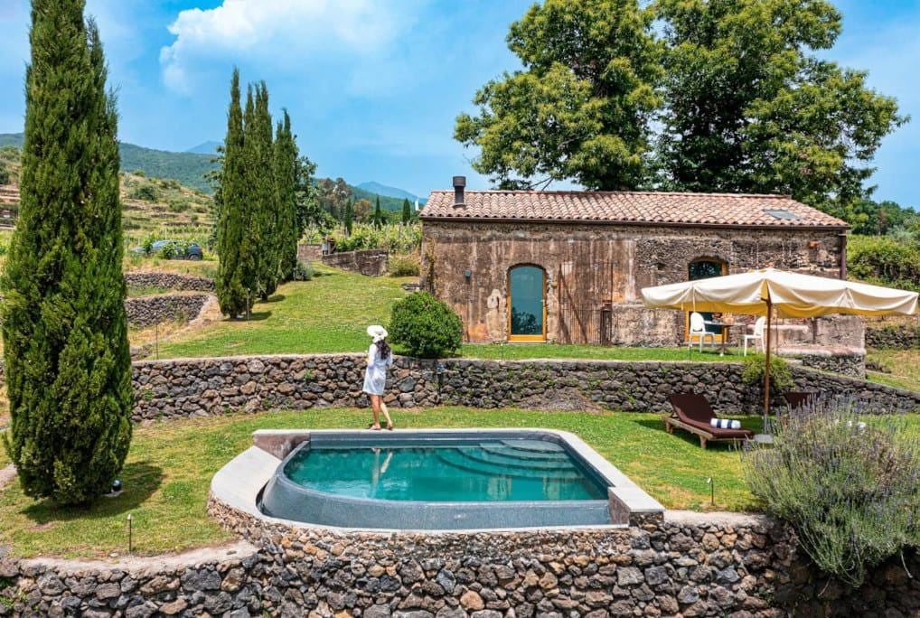 Luxurious estate suite with rustic Sicilian charm, natural wood furnishings, soft linens, and panoramic views of lush volcanic landscape at Monaci delle Terre Nere.