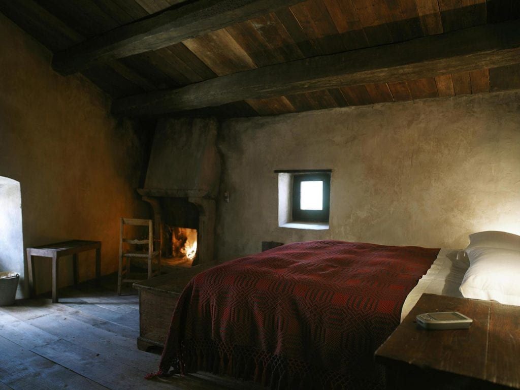 Rustic stone-walled bedroom in historic Italian hotel with soft linens, antique wooden furnishings, and warm ambient lighting in traditional mountain village setting