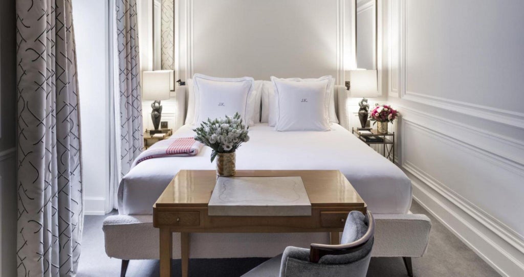 Elegant Parisian hotel room with soft cream walls, plush white bedding, classic French furnishings, and large windows illuminating luxurious interior design