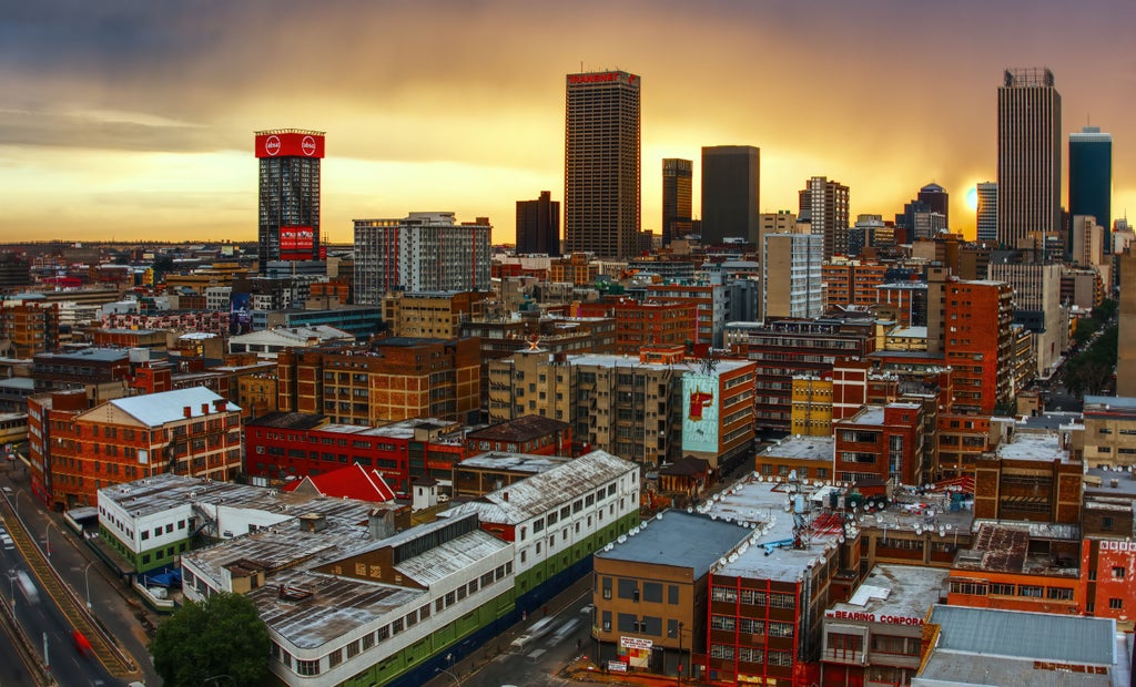 Modern luxury buildings with gleaming glass facades rise against Johannesburg's skyline at sunset, framed by lush urban greenery