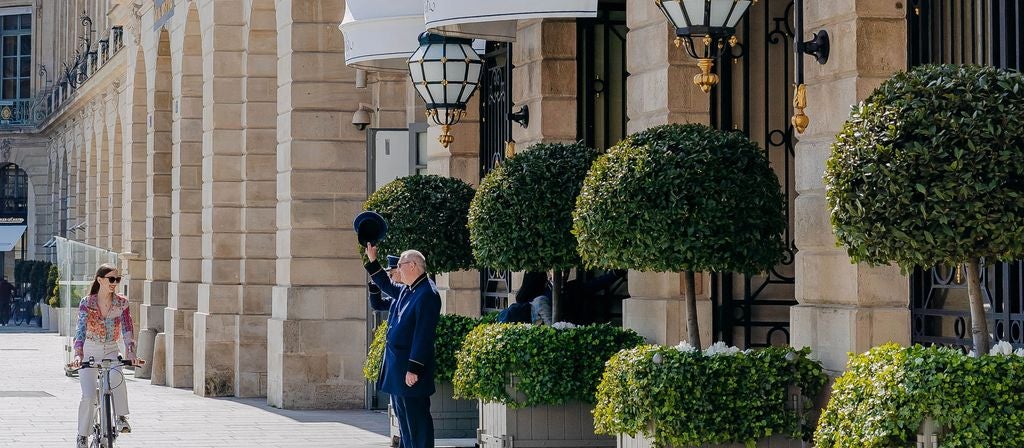 Opulent facade of Ritz Paris hotel with ornate Belle Époque architecture, gold-trimmed windows, and manicured topiaries along entrance