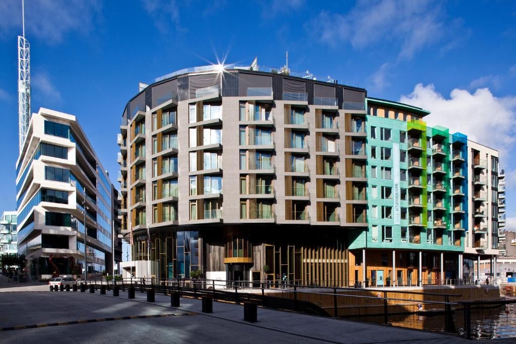 Modern luxury hotel exterior with angular glass facade, dark metal panels, and illuminated entrance reflecting against evening sky in Oslo