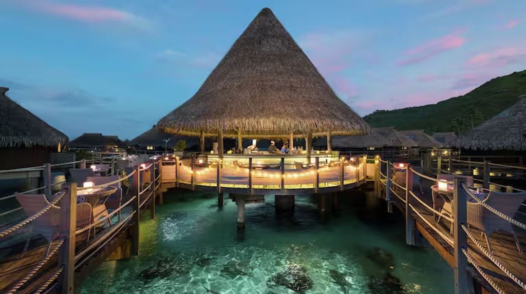 Overwater luxury bungalows with thatched roofs extending into turquoise lagoon, surrounded by lush mountains and palm trees at sunset