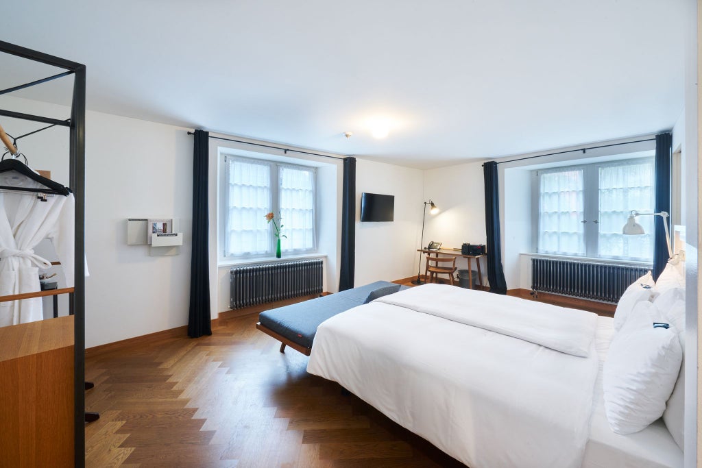 Elegant Swiss hotel room with plush white bedding, modern wood furnishings, large windows overlooking urban scenery, and sophisticated neutral color palette