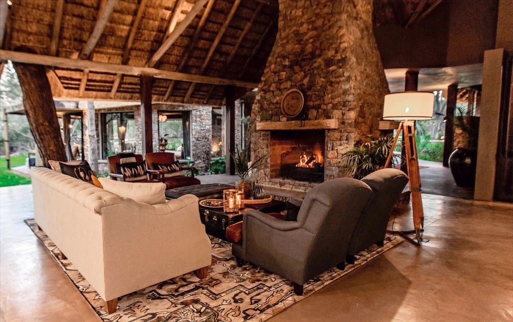 Elegant safari lodge with thatched roof and stone walls overlooking a tranquil waterhole in the South African bushveld at sunset