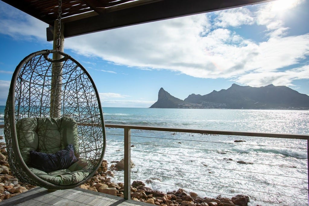 Luxury oceanfront lodge with floor-to-ceiling windows and private deck perched over rocky Atlantic coastline at sunset, Table Mountain backdrop