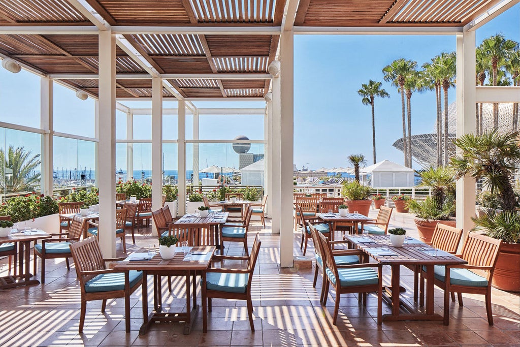 Modern luxury skyscraper hotel with distinctive blue glass facade towering over Barcelona's beachfront, palm trees line the entrance