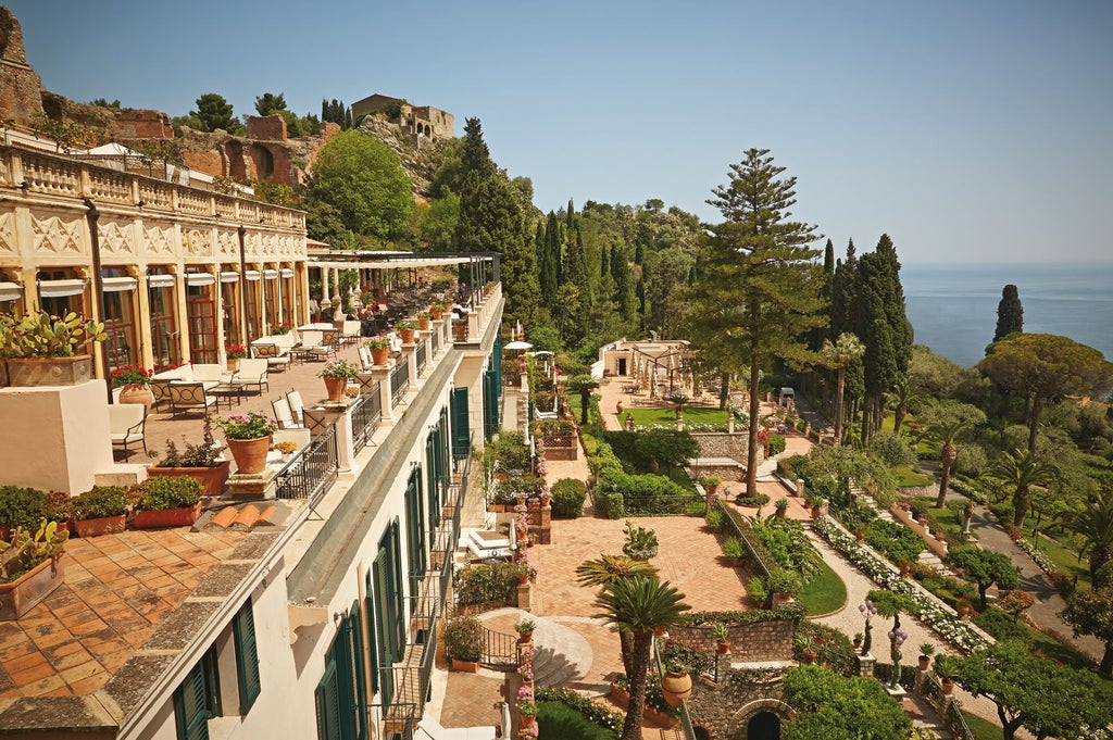 Elegant cliffside hotel with white facade and arched windows overlooking Mount Etna, set amid lush Mediterranean gardens and terraces