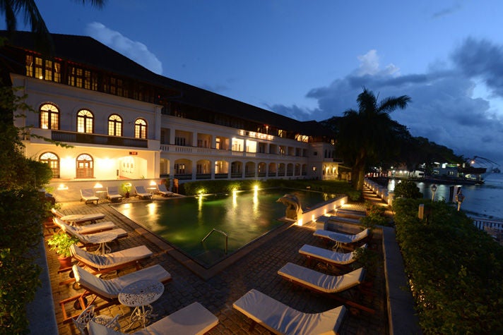 Elegant colonial-style waterfront hotel with white facade, ornate balconies, and palm trees reflecting in Kerala's backwaters at sunset