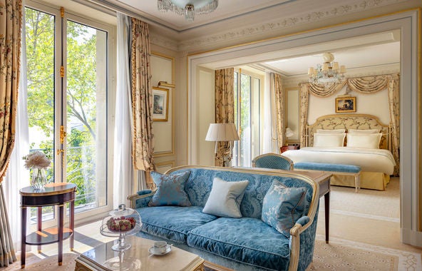 Opulent hotel room with gold-accented furnishings, crystal chandelier, Louis XV furniture, and floor-to-ceiling windows with Parisian views