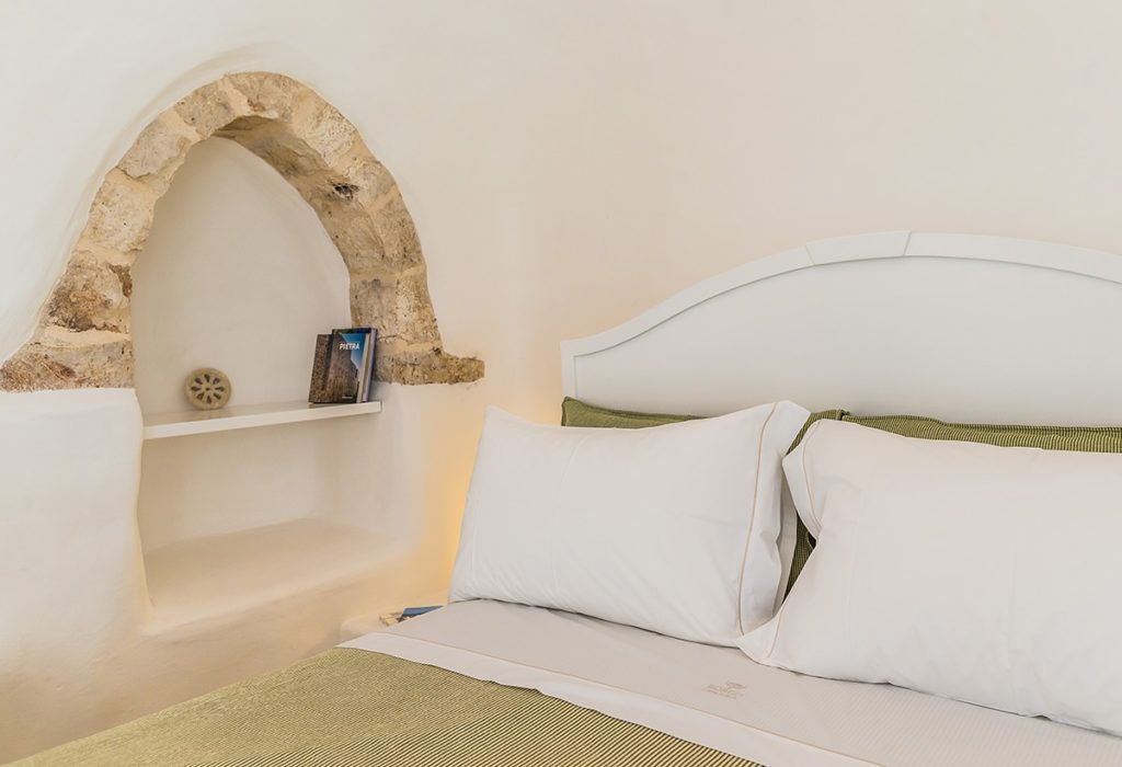 Rustic stone trulli room with white-draped bed, warm wooden accents, and soft natural light showcasing elegant minimalist design in Italian countryside