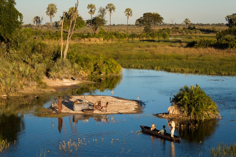 Elevated luxury safari lodge with thatched-roof suites connected by wooden walkways over wetlands at sunset in the Okavango Delta