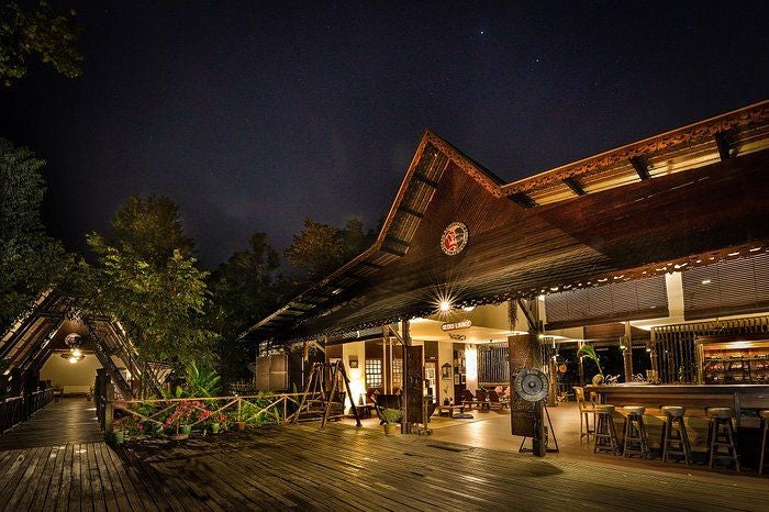 Luxurious wooden lodge with elevated walkways nestled along a river bank, surrounded by lush rainforest canopy at sunset