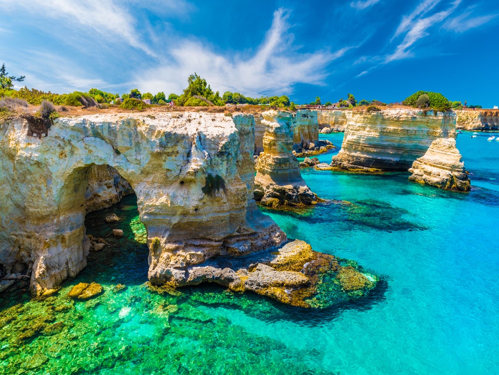 Small wooden boat gliding along crystal-clear turquoise waters of Puglia coastline, passing dramatic limestone cliffs and hidden caves