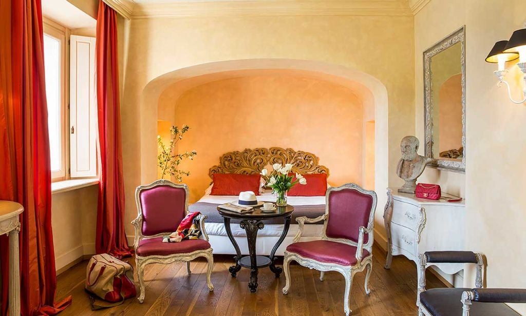 Elegant deluxe hotel room with plush king bed, soft neutral tones, rustic wooden headboard, and expansive Tuscan landscape view through large windows.