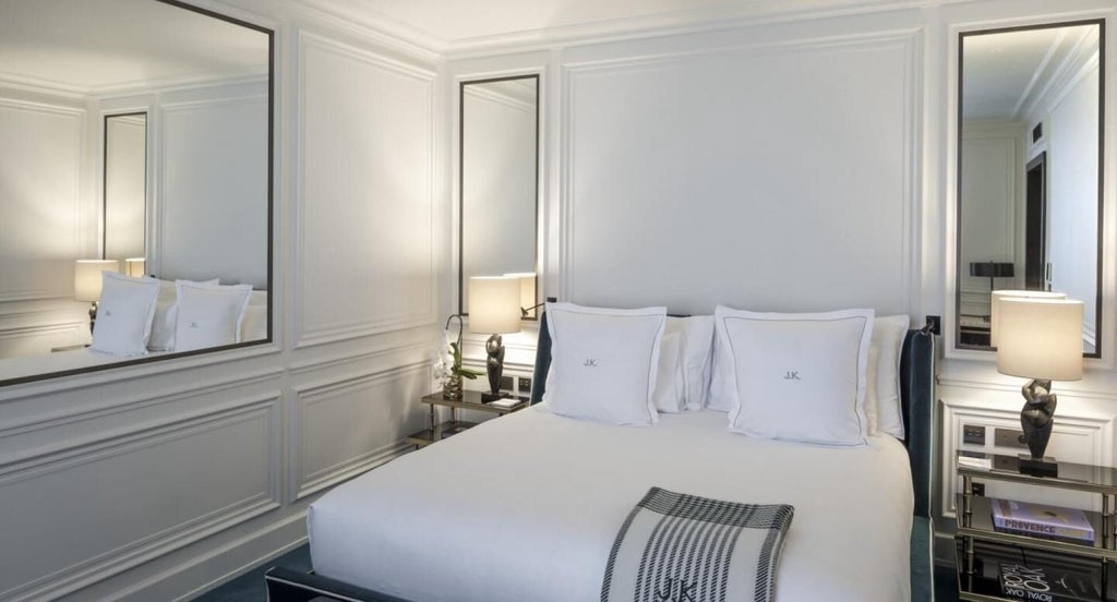 Elegant Parisian hotel room with plush white bedding, soft neutral tones, designer furnishings, and large windows overlooking a chic urban landscape