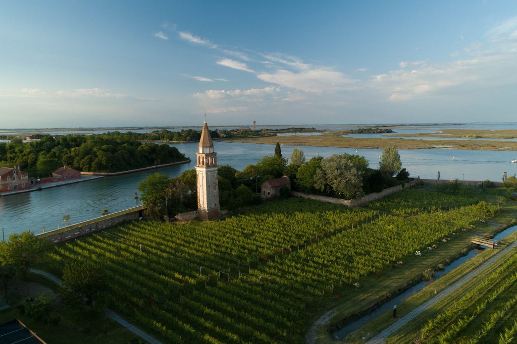 Elegant Venissa Wine Resort's facade reflects in calm lagoon waters, with lush gardens and historic stone architecture at sunset