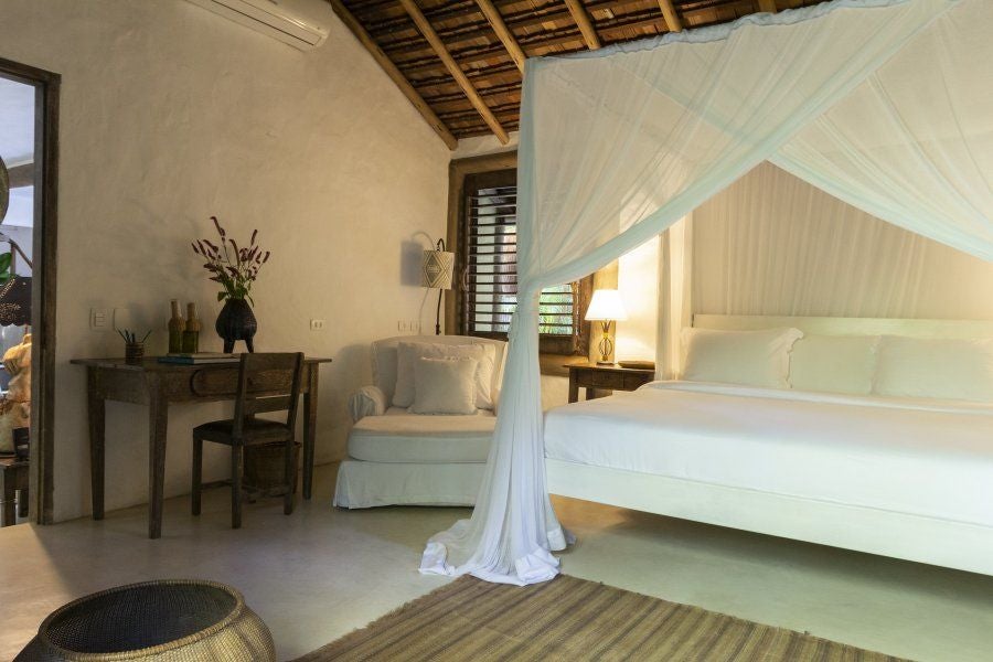 Luxurious Brazilian hotel room with rustic wooden furniture, white linens, and a glimpse of lush tropical greenery through large windows at UXUA Casa Hotel and Spa