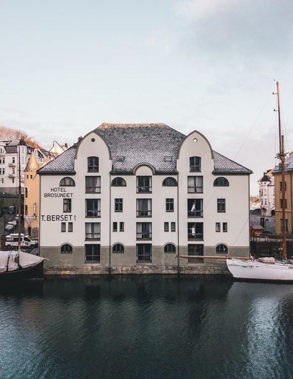 Historic waterfront hotel with sleek wooden facade, glass windows, and moody Nordic lighting reflecting on calm harbor waters at dusk