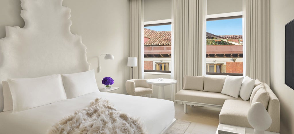 Elegant hotel room with floor-to-ceiling windows, king bed, modern furnishings, and panoramic Madrid cityscape views at golden hour