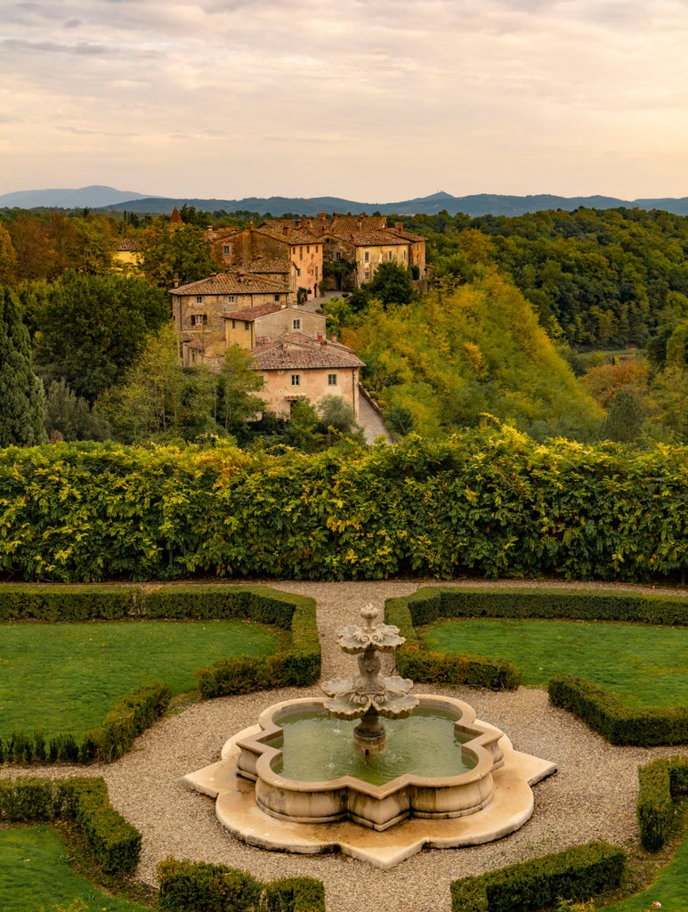 Historic stone villa surrounded by manicured Tuscan gardens and cypress trees, with elegant terrace overlooking rolling vineyard hills at sunset