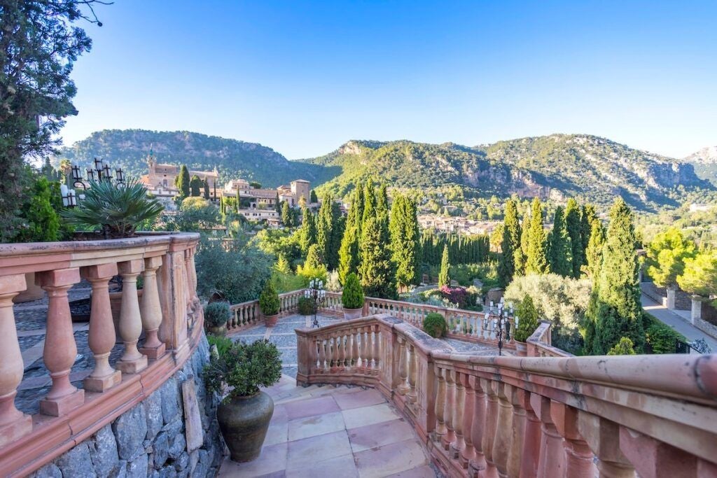 Elegant stone hotel with arched windows nestled in lush Mallorca hills, terrace seating overlooks Mediterranean gardens at sunset