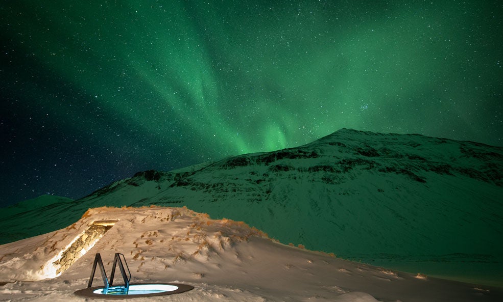 Vibrant green and blue Aurora Borealis dancing across dark Icelandic winter sky, with snow-covered mountains and a luxurious glass-domed observation cabin in foreground.