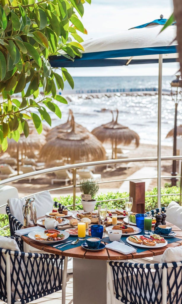 Luxurious seaside resort with Mediterranean-style architecture, white buildings, palm trees, and blue swimming pool overlooking pristine sandy beach in Spain