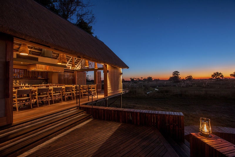 Elevated wooden safari lodge with private deck overlooking Okavango Delta wetlands, featuring thatched roof and luxury outdoor seating