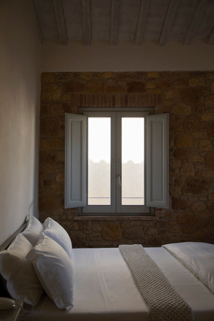 Elegant minimalist hotel room with stone walls, crisp white linens, rustic wooden furnishings, and large windows overlooking Tuscan countryside landscape