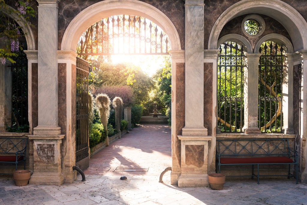 Elegant Italian palazzo with ornate stone facade, grand arched windows, wrought-iron balconies, and lush garden courtyard in warm sunlight