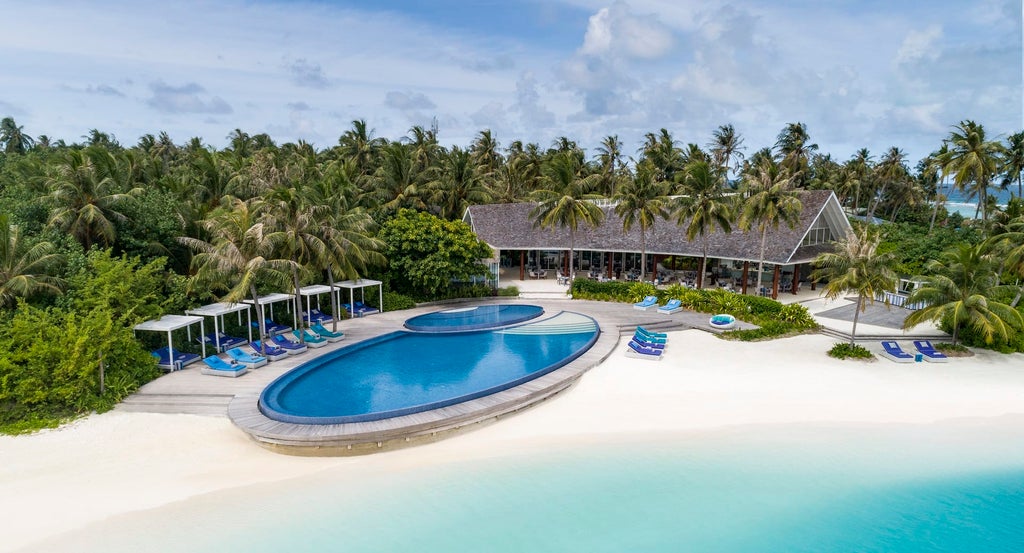 Modern overwater villa with infinity pool merging into turquoise Maldivian lagoon, surrounded by wooden deck and tropical palms.